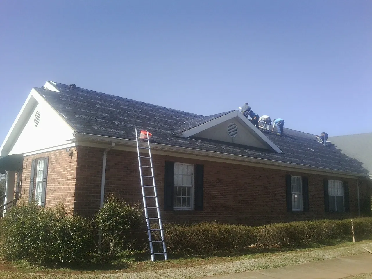Crew tearing shingles off a brick ranch roof with a ladder set — Emergency Roof Repair work by our crew in East Jewett