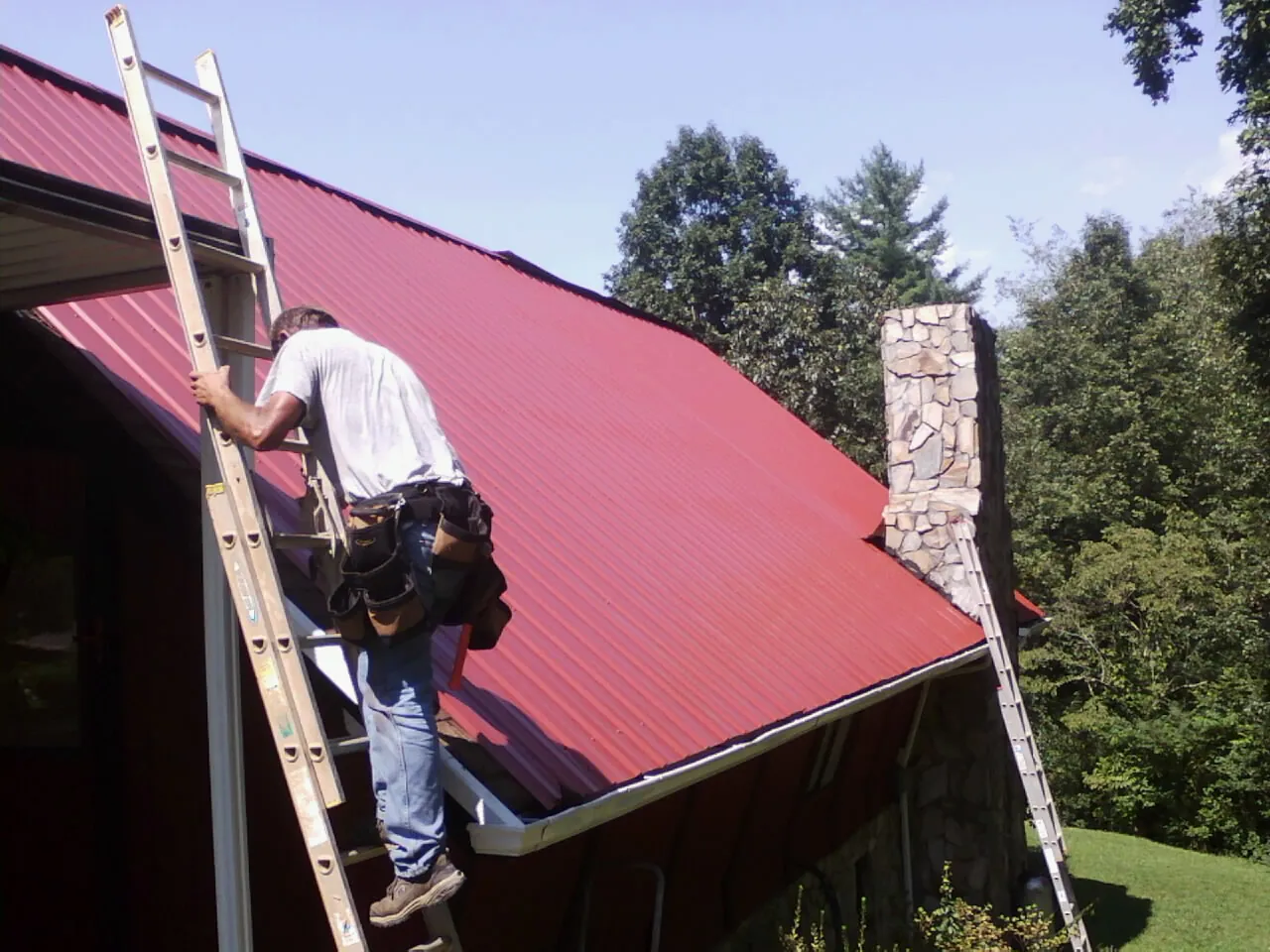 Roofer on a ladder beside a red metal roof and stone chimney — Soffit & Fascia Repair by Roofing Brothers in East Jewett